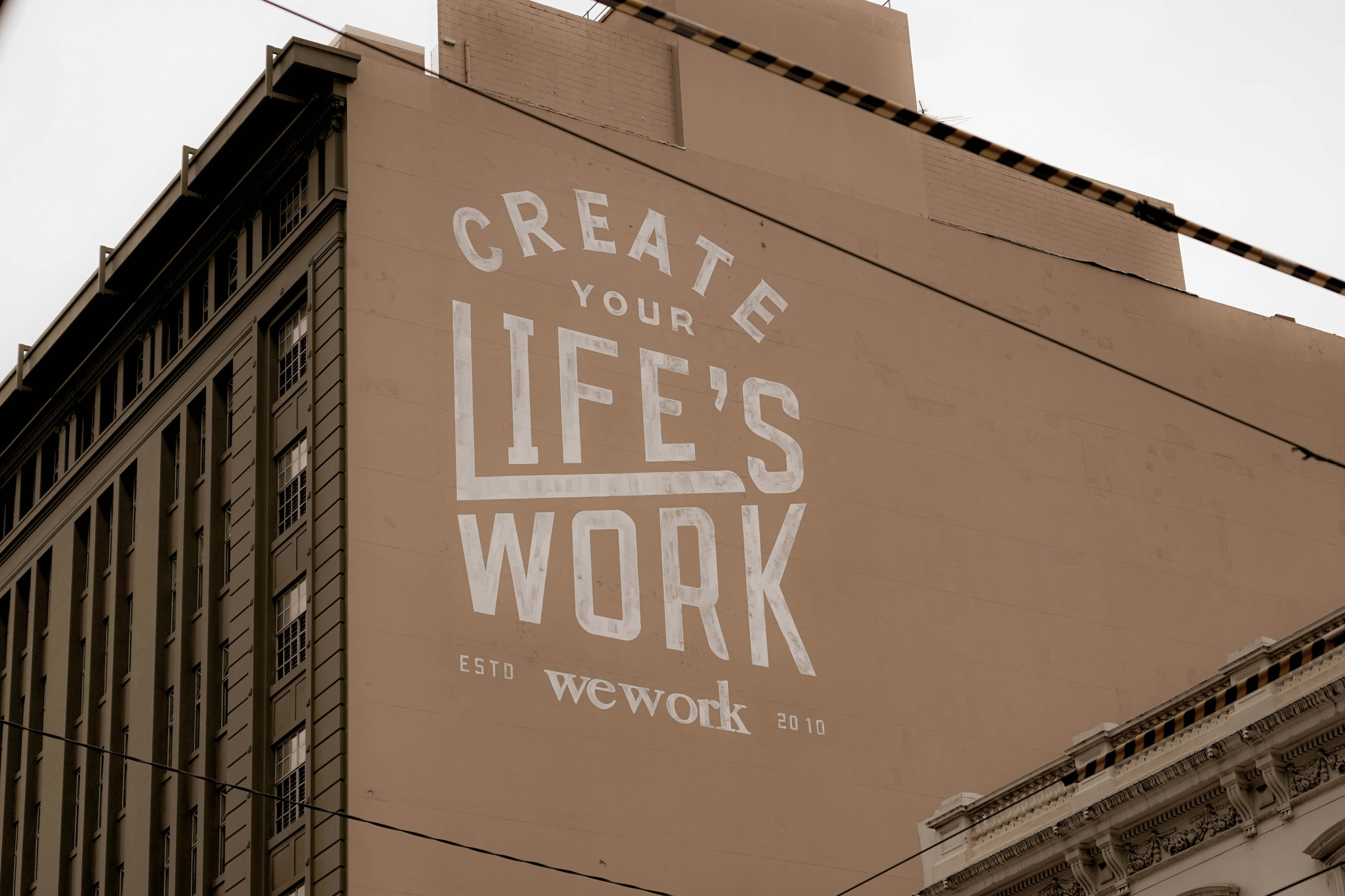 A large building with a beige wall featuring a white mural that reads 'CREATE YOUR LIFE'S WORK' and 'wework ESTD 2010'. Another building with many windows is visible on the left.