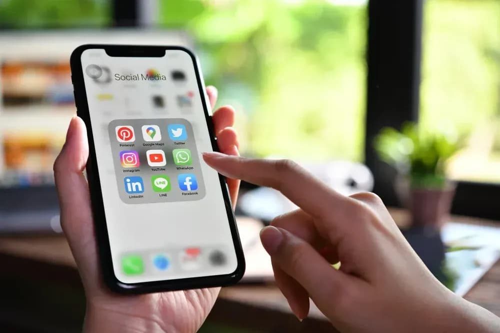Person holding a smartphone displaying a 'Social Media' folder with app icons like Instagram, Facebook, YouTube, and WhatsApp.
