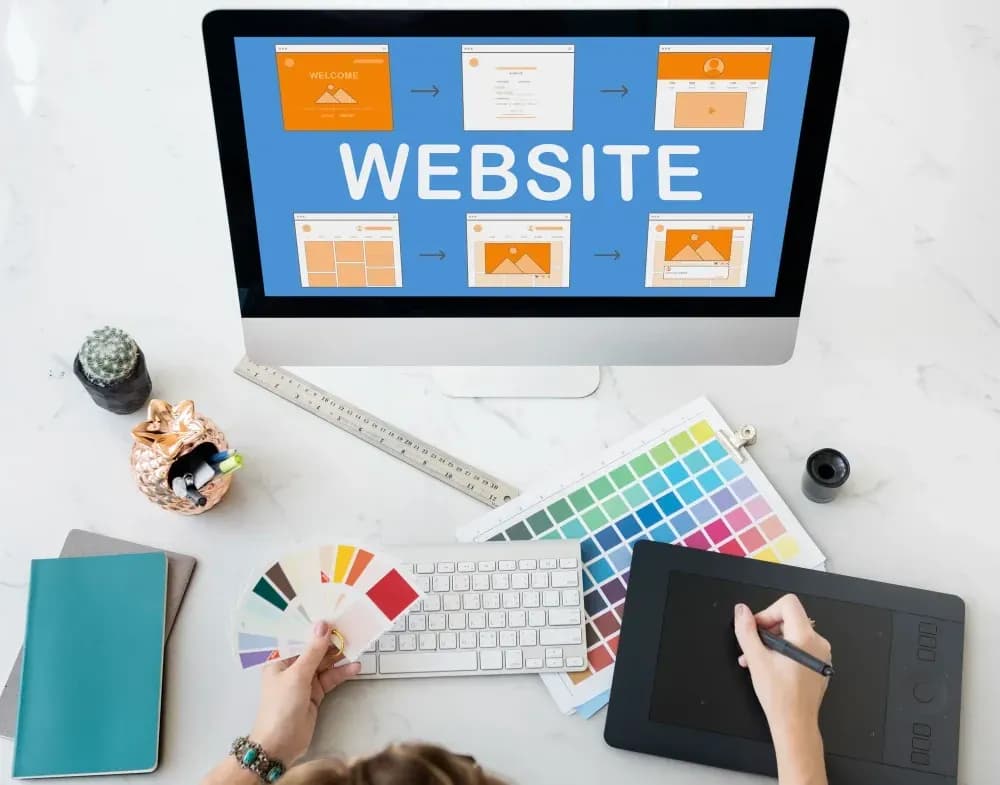 Overhead view of a designer creating a website, showing UI/UX wireframes on a monitor, color palettes, and a drawing tablet.