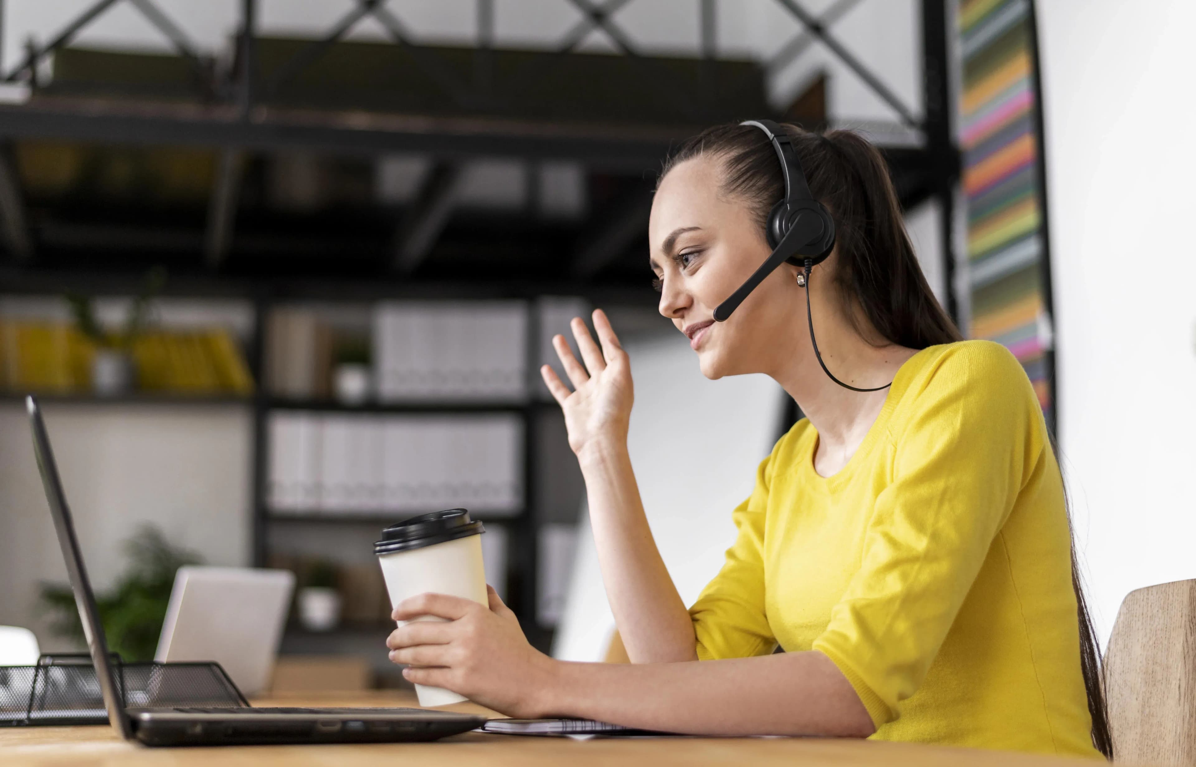 Young woman in yellow shirt with headset on video call, holding coffee cup, gesturing at laptop in home office.
