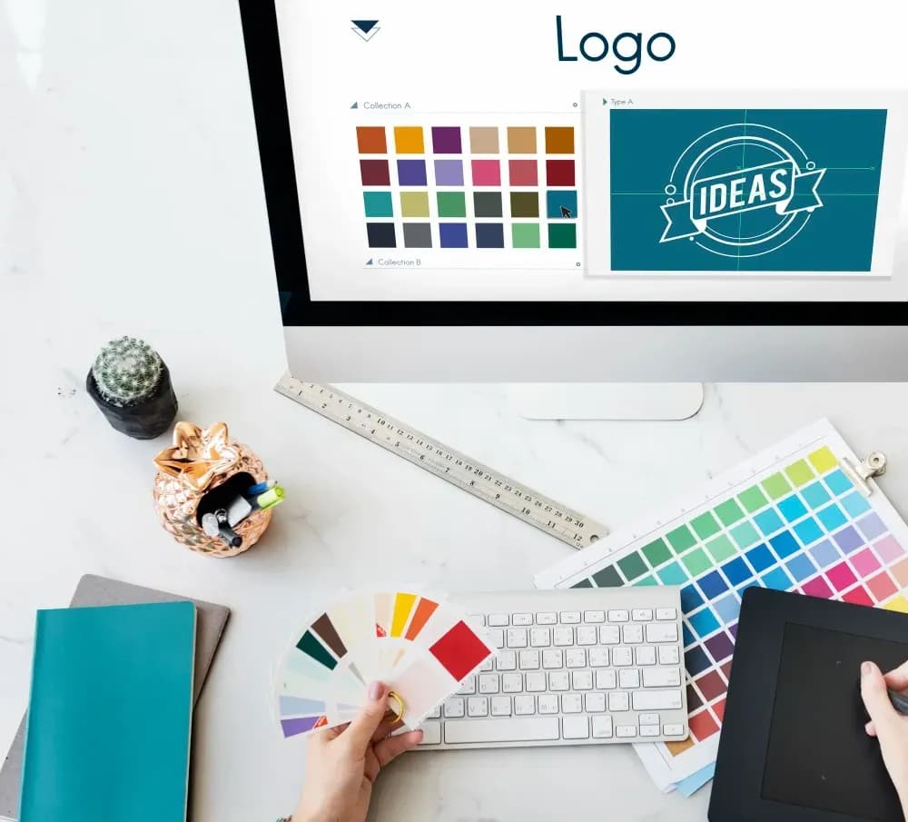 Overhead view of a graphic designer's desk with a computer displaying logo ideas and color palettes. Hands hold a color swatch book, surrounded by a keyboard, ruler, and graphics tablet.