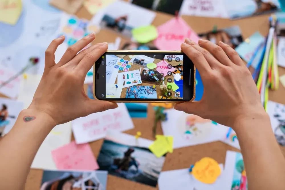 Person taking a photo of a colorful vision board with a smartphone, capturing travel photos, notes, and creative ideas.