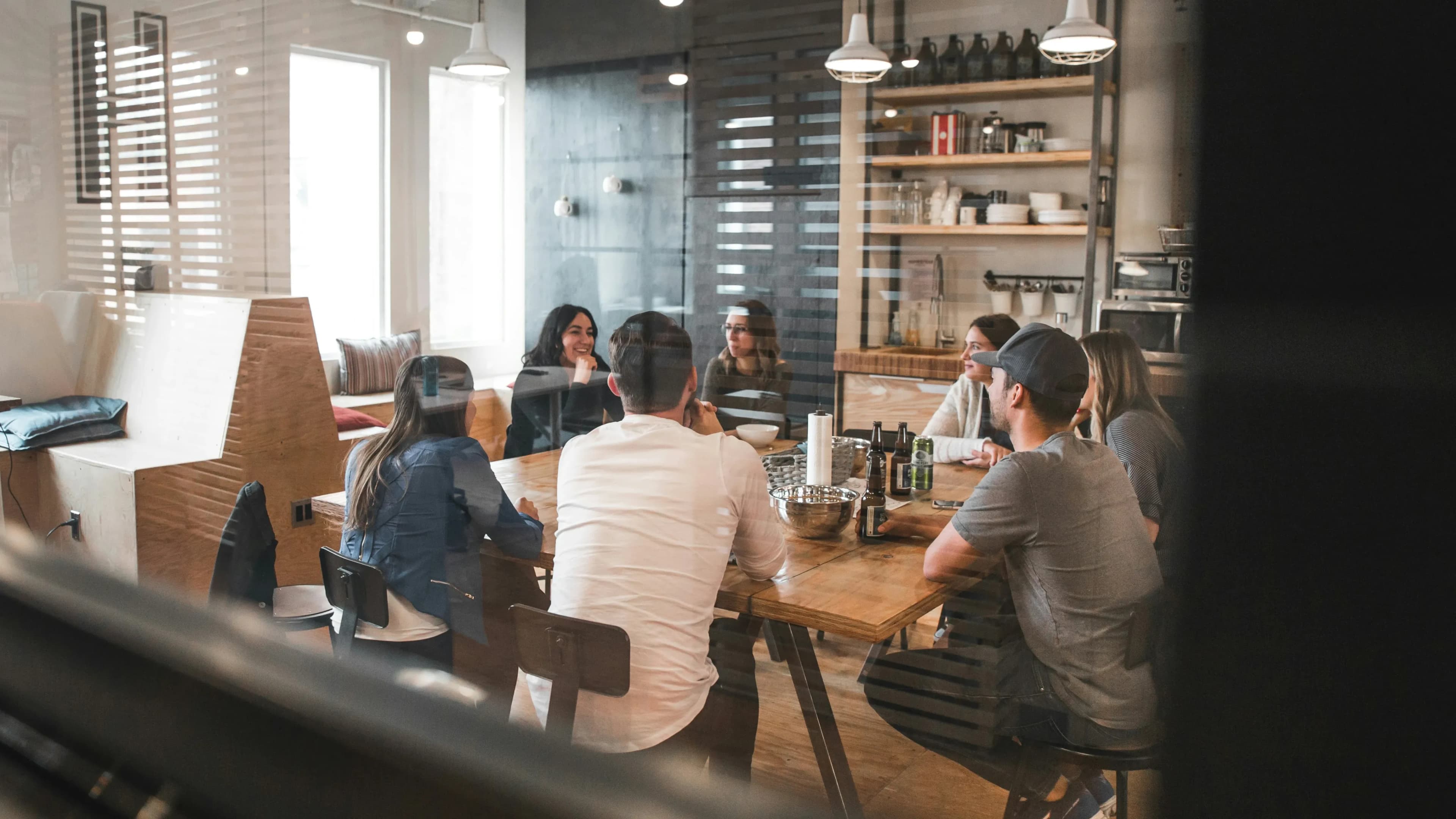 A diverse group of professionals collaborating around a wooden table in a modern, bright office space with drinks and snacks.