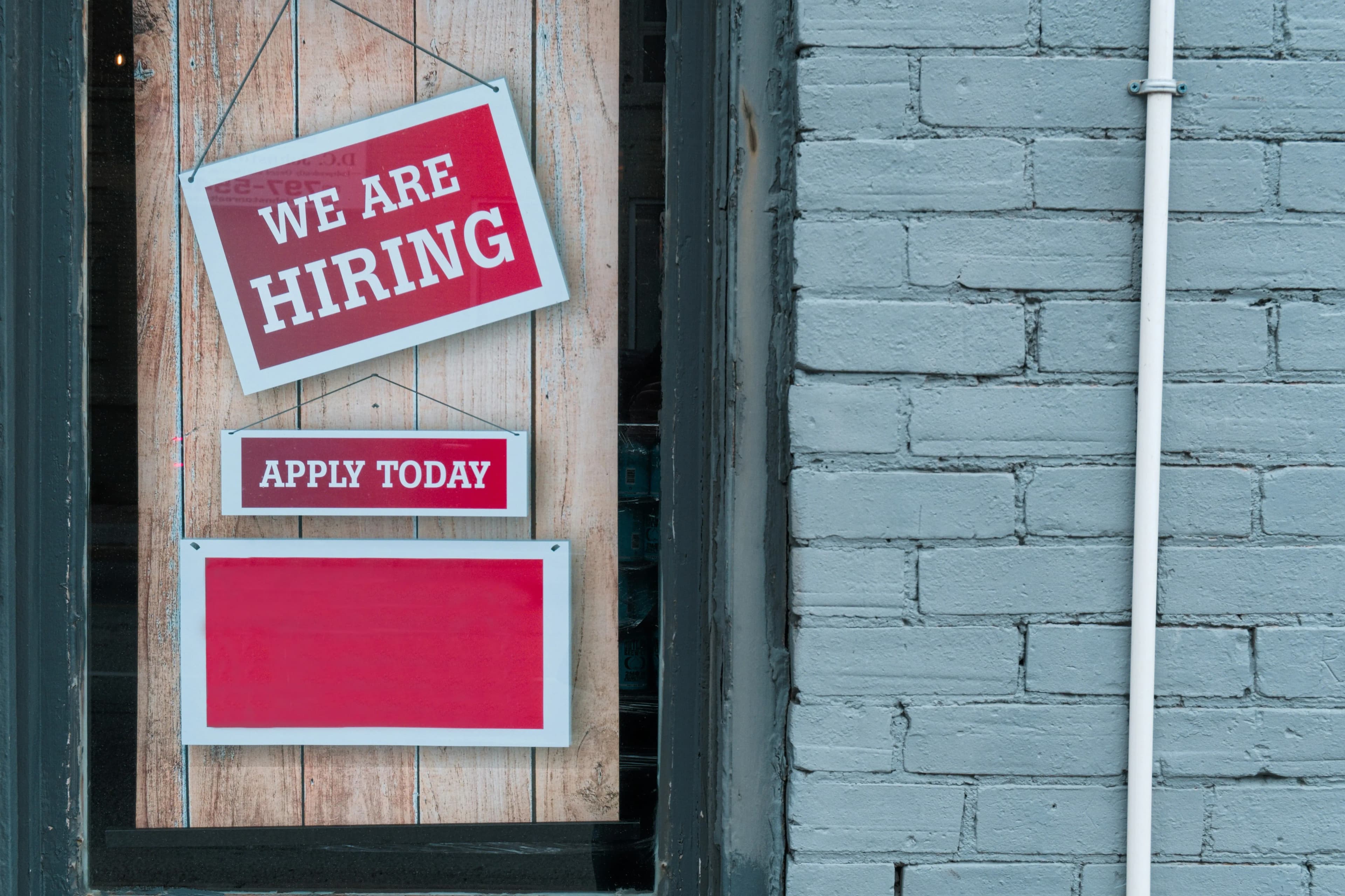 Red 'WE ARE HIRING' and 'APPLY TODAY' signs displayed on a rustic wooden door, beside a light blue brick wall.
